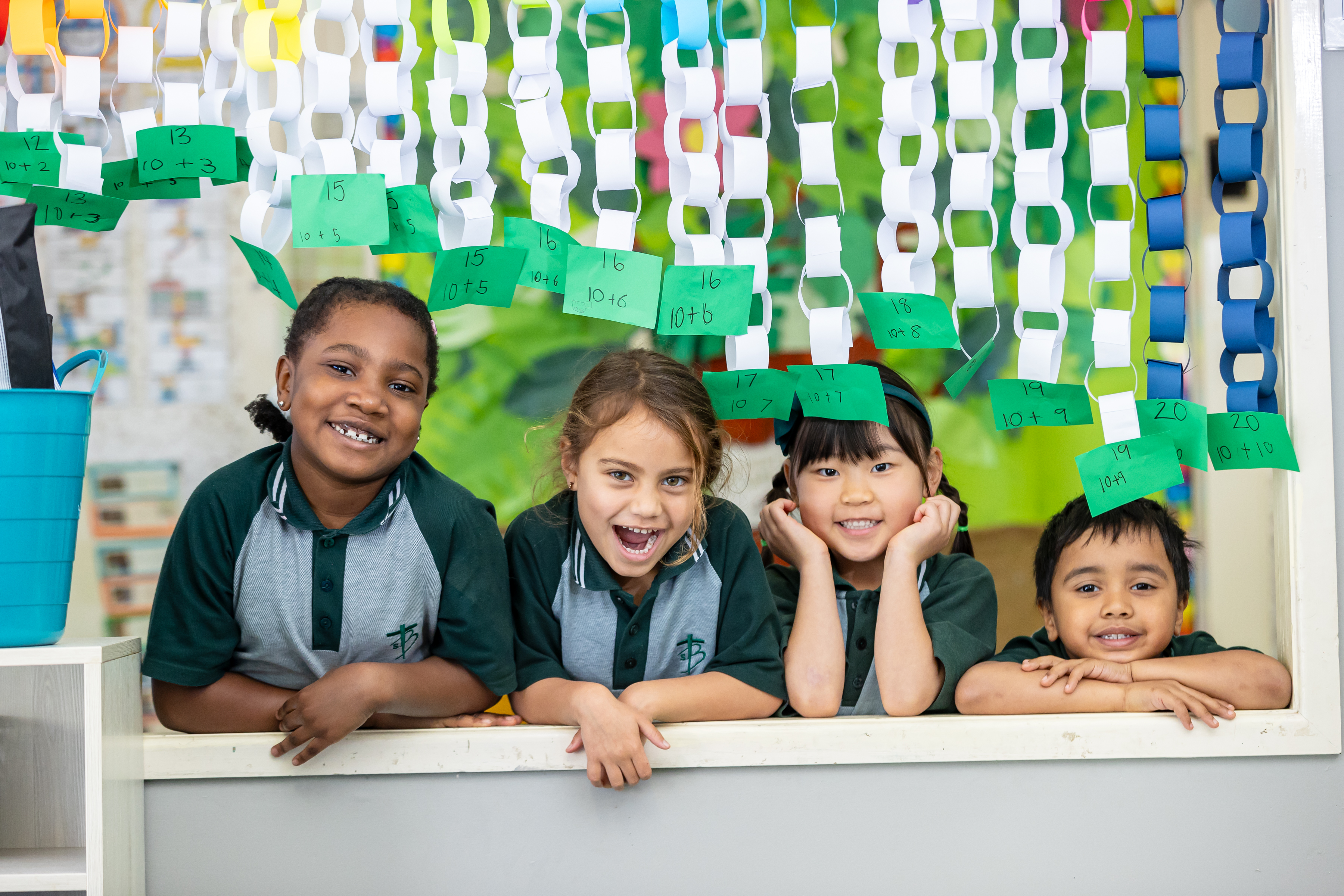 Four children leaning on a classroom ledge beneath hanging paper chains, with green cards displaying addition sums above them.